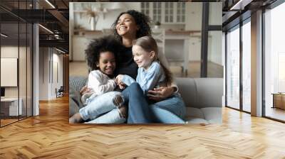 Overjoyed young African American mother play with little multiracial daughters at home, happy biracial mom sit on couch tickle have fun with smiling small multiethnic girls kids on weekend together Wall mural