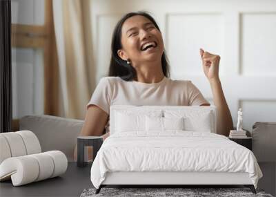 Overjoyed young Asian woman holding phone, celebrating success, showing yes gesture, sitting on cozy couch at home, excited laughing girl received good news, great shopping offer, online lottery Wall mural