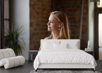 Peaceful thoughtful long haired young woman in casual looking at window away with folded hands, standing in loft space home, thinking, dreaming, smiling at good thoughts, pondering on future plans Wall mural