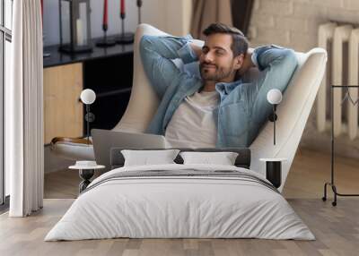 Peaceful young man daydreaming in comfortable armchair, holding computer. Wall mural