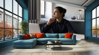 Pensive millennial Indian female student busy thinking studying online on laptop at home. Thoughtful young ethnic woman make plan or decide working distant on computer, writing or listing in notebook. Wall mural