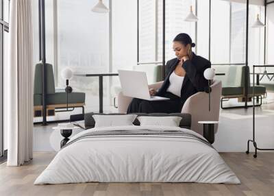 Pensive young African American businesswoman in formal suit typing on laptop in modern co-working space, sitting in comfortable armchair with computer on lap, thinking on project Wall mural