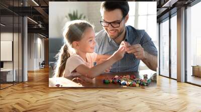 Playful young Caucasian father and little happy daughter have fun making bracelets accessories at home together. Loving dad and small girl child engaged in funny hobby creative activity string beads. Wall mural