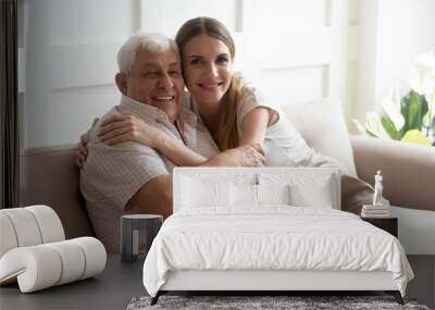 Portrait happy older father and adult daughter hugging, looking at camera, posing for photo, beautiful young woman with smiling mature dad or grandfather sitting on cozy sofa at home, two generations Wall mural