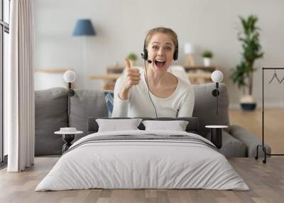 Portrait of excited young female wearing headset show thumbs up to camera recommending online course or training, happy girl in earphones sit with laptop at home gesture liking educational program Wall mural