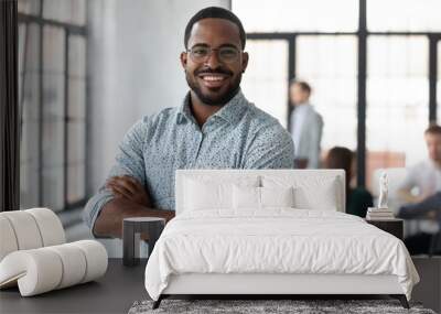 Portrait of happy African American small business owner posing with hands folded. Millennial black male team leader smiling, looking at camera, employees working in modern office behind. Head shot Wall mural