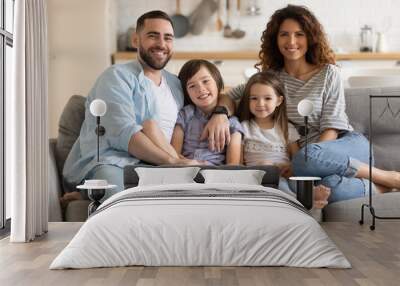 Portrait of happy married couple embracing little kids son daughter, sitting together on comfortable couch in modern studio living room. Smiling cheerful spouses posing for photo with children. Wall mural