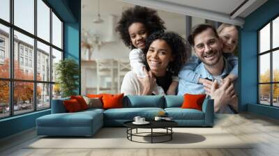 Portrait of happy multiracial couple enjoying sweet family moment with adorable little mixed raced daughters at home. Smiling cute small stepsisters cuddling cheerful parents, looking at camera. Wall mural