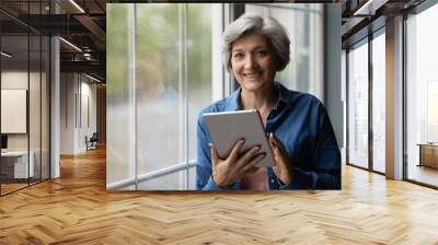 Portrait of smiling elderly female with tablet device. Middle-aged woman looking at camera pose near window holding modern electronic gadget, easy comfort remote services for older generation concept Wall mural