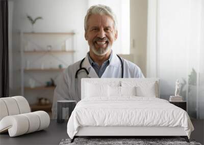 Portrait of smiling middle-aged Caucasian male doctor in white medical uniform use modern smartphone in clinic. Happy mature man GP or therapist look at camera consult patient on cellphone online. Wall mural