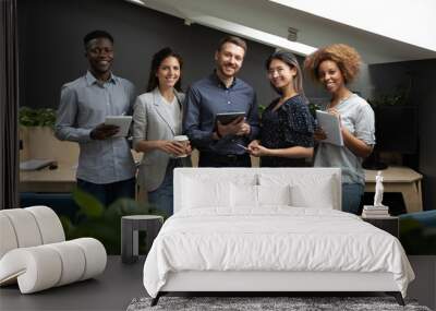 Portrait of successful diverse employees team holding electronic devices, looking at camera, happy smiling colleagues standing in modern office, posing for photo, motivated for business success Wall mural