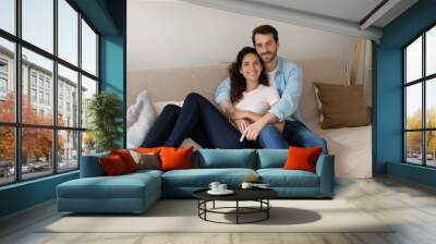 Portrait of young wife and husband hugging, sitting on couch, smiling attractive woman and man cuddling, looking at camera, posing for family photo at home, relaxing on sofa, enjoying lazy weekend Wall mural