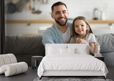 Portrait smiling father and little daughter hugging, looking at camera, sitting on cozy couch at home, happy young dad and cute adorable girl posing for family photo, enjoying leisure time together Wall mural