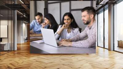 Positive diverse couple of office colleagues engaged in online presentation on laptop working together at large desk in office co-working space, using computer, watching work content smiling Wall mural