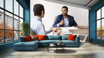 Positive handsome young Indian businessman talking to male colleague at meeting table, smiling. Business partners discussing investment, collaboration, teamwork, startup marketing strategy Wall mural