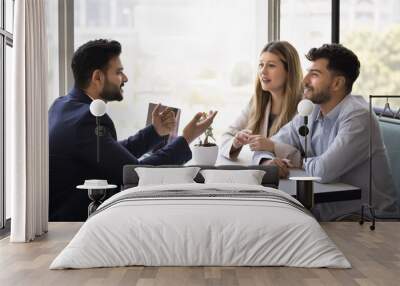 Positive married couple consulting financial expert, real estate agent at meeting, listening to young Indian business consultant in formal suit, sitting at table with paper agreement Wall mural