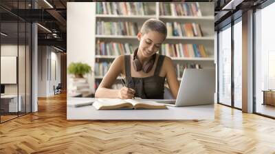 Positive student girl studying in campus library, writing notes, listening to webinar, online lesson on laptop, doing academic research study, enjoying education, knowledge Wall mural