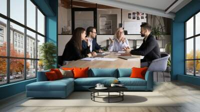 Senior company owner woman talking to team of younger managers on corporate meeting, sitting at boss place at large table, explaining teamwork strategy, smiling, discussing collaboration Wall mural