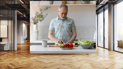 Senior grey haired vegetarian man cooking lunch in kitchen, cutting fresh vegetables at table, preparing organic meal, keeping healthy diet, caring of clean nutrition. Culinary, eating at home concept Wall mural