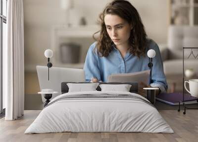 Serious busy young freelancer girl checking financial papers, legal documents at work table, using at laptop, looking at display, reviewing contract, agreement, consulting online paperwork service Wall mural