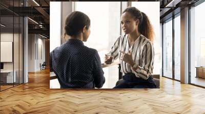 Serious pretty young African employee girl talking to colleague, office friend. Diverse project coworkers discussing project strategy, teamwork tasks, talking at large window in hall Wall mural