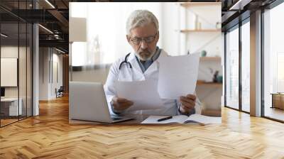 Serious professional senior elderly doctor doing paperwork checking medical documents at workplace. Concentrated old physician reading medic form analyzing patient diagnosis or report in hospital. Wall mural