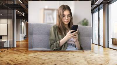 Serious teenager girl using cellphone at home, watching videos on streaming movies and TV platforms, playing mobile games, chatting with friends through messenger. Modern gadgets overuse, bad habit Wall mural