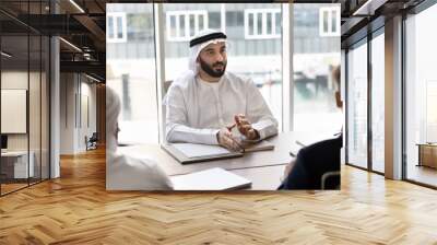 Serious young handsome Arab entrepreneur in traditional clothes talking to diverse partners at meeting, sitting at large table, speaking, discussing teamwork, cooperation, contract terms Wall mural