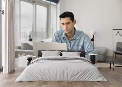 Serious young Hispanic freelance entrepreneur working at computer, sitting at home workplace desk, typing on laptop, sitting at table in cozy apartment, using gadget for online business communication Wall mural