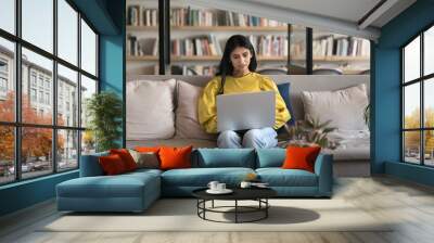 Serious young Indian entrepreneur woman typing on laptop in modern co-working space, sitting on couch, using computer. College student working on research study project in library Wall mural