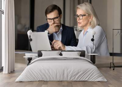 Serious younger and elder professional colleagues in glasses working together at laptop. Mature project manager, businesswoman showing online presentation on laptop to partner man Wall mural