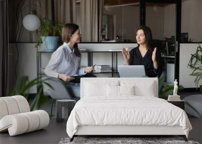 Sharing experience. Capable young indian female mentor explaining student intern corporate work principles using laptop and paper documents, two diverse women colleagues discussing business project Wall mural