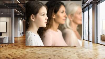 Side shot of serious little tween child girl standing in row with mom and grandmother in blurred background, looking forward away, posing for family portrait of three female generations Wall mural