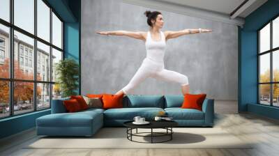 Side view portrait of beautiful young woman wearing white tank top working out against grey wall, doing yoga or pilates exercise. Standing in Warrior two pose, Virabhadrasana. Full length Wall mural