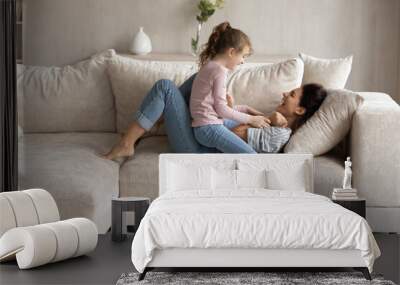 Small ethnic girl child and excited young Latino mom relax on couch in living room feel playful on weekend together. Overjoyed Hispanic mother and little daughter have fun play and tickle at home. Wall mural