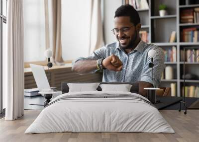 Smiling African American businessman wearing glasses checking time, looking at wristwatch on hand, positive happy young man entrepreneur sitting at desk, planning work, task management concept Wall mural