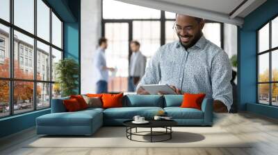 Smiling African American male employee use modern tablet gadget with wireless Internet connection at workplace, happy confident biracial man worker texting or messaging on Pad device in office Wall mural