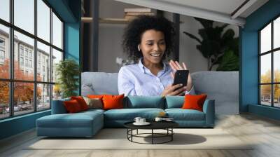 Smiling African American woman in wireless earphones waving hand at phone camera, holding smartphone, happy young female chatting with friends online by video call at home, blogger greeting viewers Wall mural