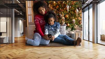 Smiling African American woman with adorable daughter using phone, sitting on warm floor near Christmas tree at home, happy mother and little girl holding smartphone, shopping online, choosing gifts Wall mural