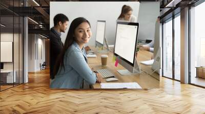 Smiling asian office employee looking at camera working with multiracial colleagues, happy young company manager sitting at corporate workplace, cheerful team member posing with desktop computer Wall mural