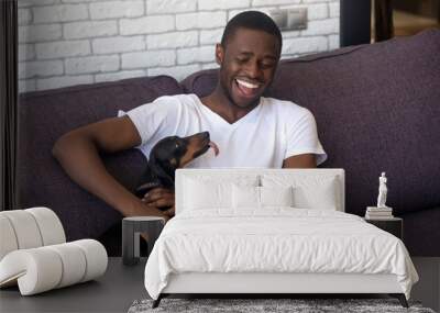 Smiling black guy relax on sofa playing with dackel dog Wall mural