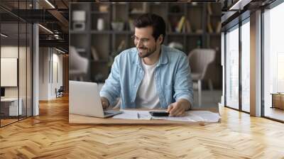 Smiling businessman in glasses using laptop, calculating bills, managing finances, happy young man planning budget, sitting at desk at home, browsing online banking service, satisfied by money refund Wall mural