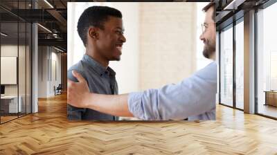 Smiling Caucasian businessman handshake tap shoulder of excited African American male employee greet with promotion, happy diverse businesspeople talk laugh close deal at meeting in office Wall mural