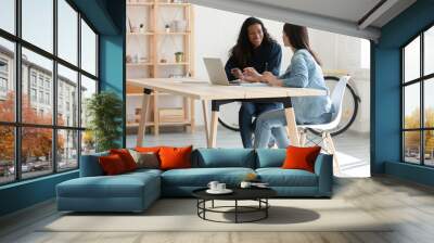 smiling female colleagues sit at desk in office talk brainstorm work together on laptop, happy woman Wall mural