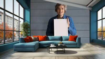 Smiling grey haired senior lady head shot. Positive 60s mature adult woman holding blank empty white picture frame, standing at studio background, looking at camera. Female portrait Wall mural