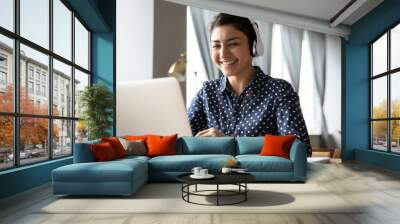 Smiling Indian girl wearing headphones using laptop, looking at screen, happy young female listening to favorite music while working online on project, excited student learning language Wall mural