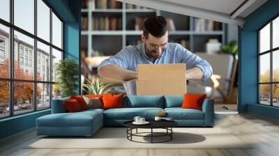 Smiling man sitting at workplace home office desk wearing glasses unpacking opening cardboard box long awaited parcel with goods bought on-line. Satisfied client of shipping delivery service concept Wall mural