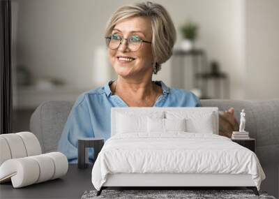 Smiling mature woman looking away while holding tablet seated on sofa at home, older generation and modern wireless technology and internet usage for online shopping, communication on carefree pastime Wall mural