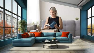 Smiling mature woman sitting on sofa unpacking box, get package, opening received parcel with ordered books using easy and comfortable on-line bookshop delivery services. Hobby, leisure, book lover Wall mural