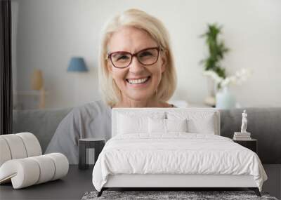 Smiling middle aged mature grey haired woman looking at camera, happy old lady in glasses posing at home indoor, positive single senior retired female sitting on sofa in living room headshot portrait Wall mural